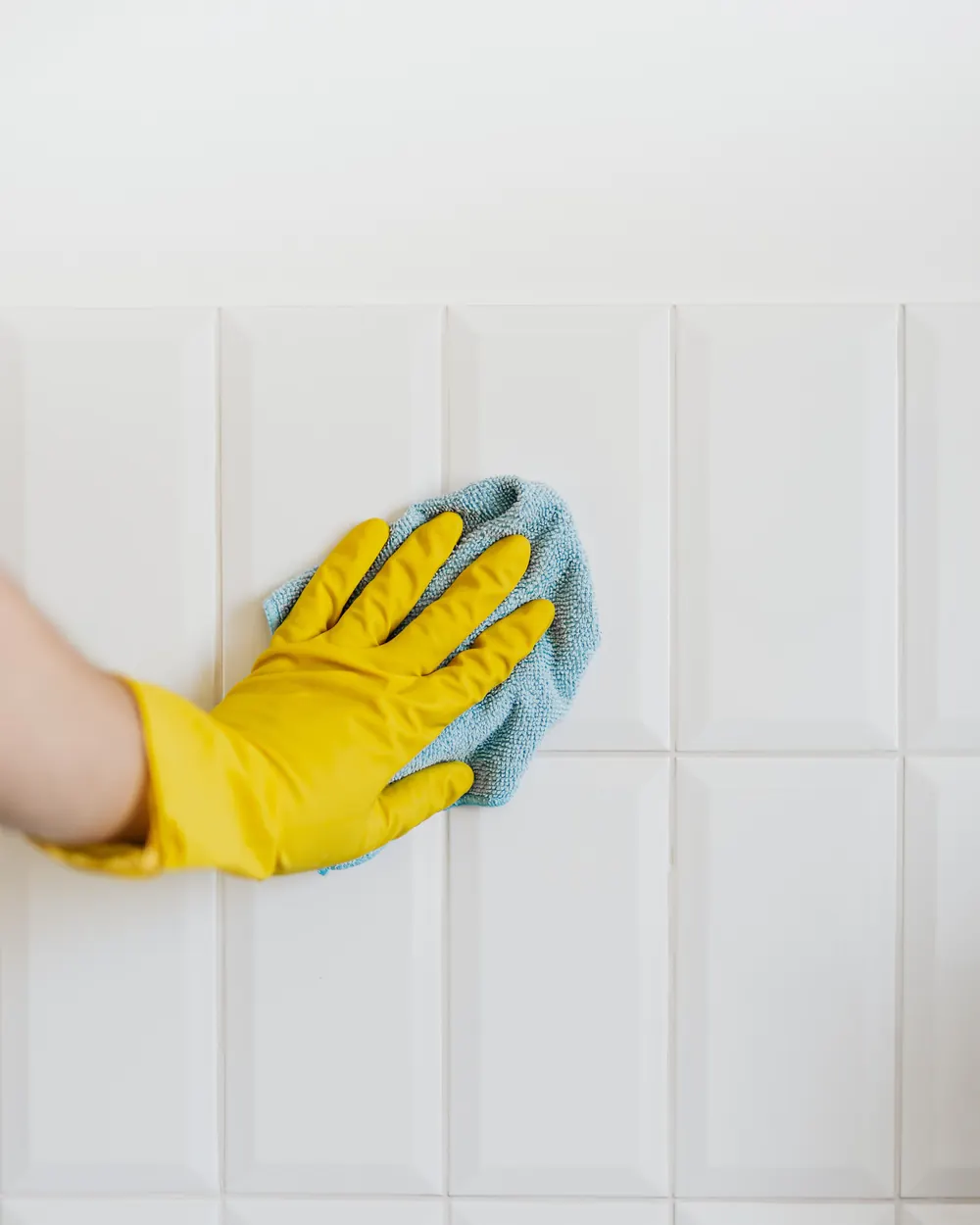 Yellow gloved hand wiping white bathroom wall tiles